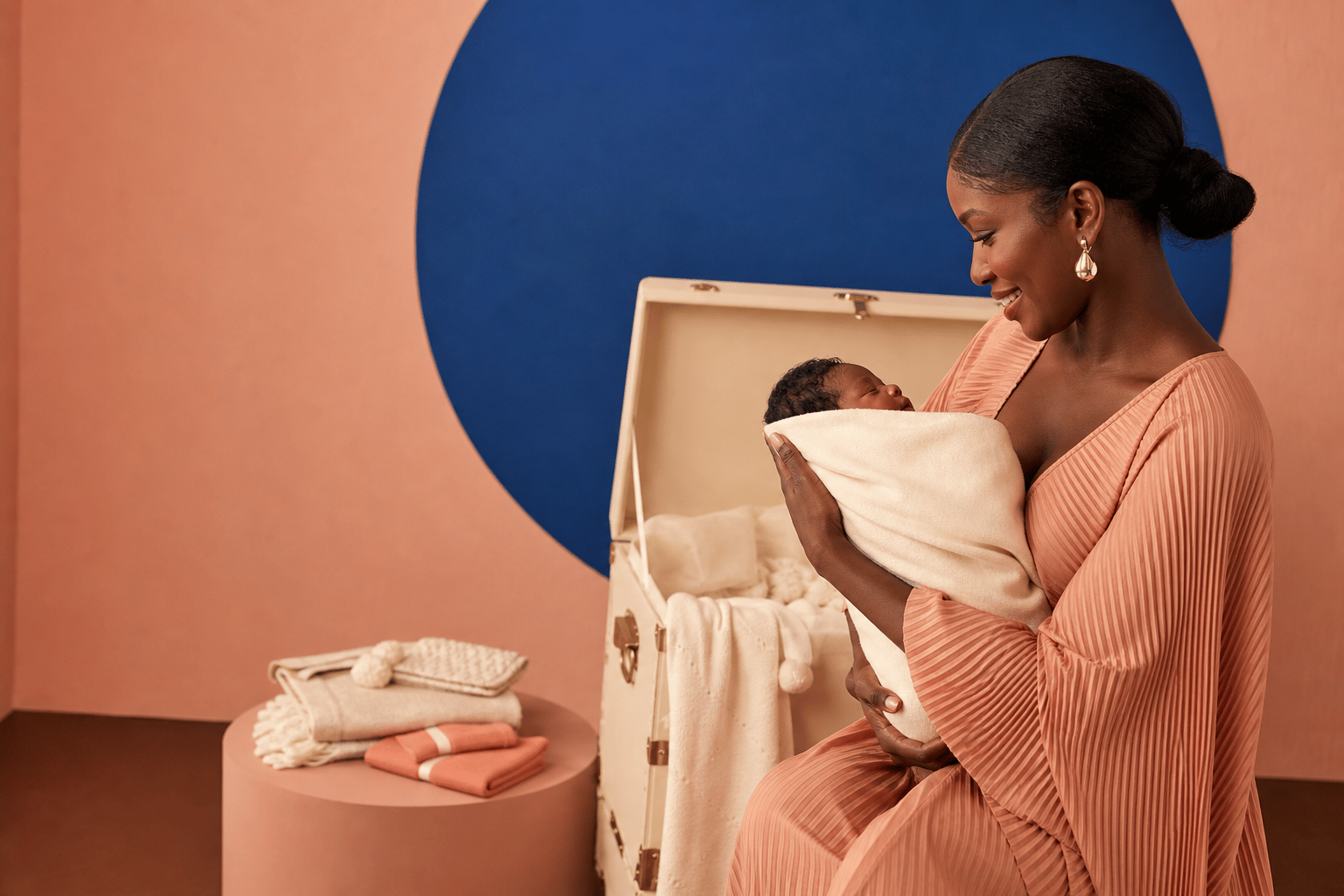 A mother in a terracotta dress cradling her newborn beside an open Treasured Trunks keepsake trunk