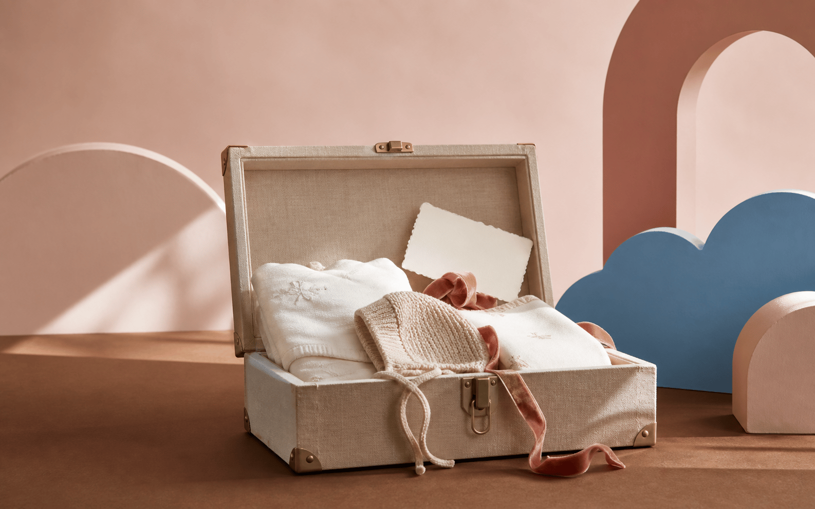 An open ivory keepsake trunk lined in linen, holding folded newborn essentials and a velvet ribbon